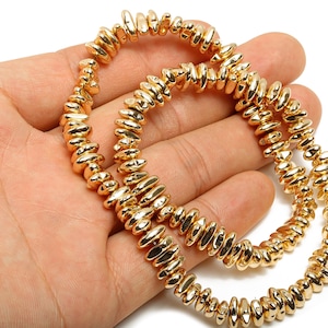 Può includere: Due fili di gioielli con perline color oro. Le perline sono di forma ovale e hanno una finitura lucida. I gioielli sono esposti sulla mano aperta di una persona su sfondo bianco. Le perline sono lunghe circa 0,6 cm.