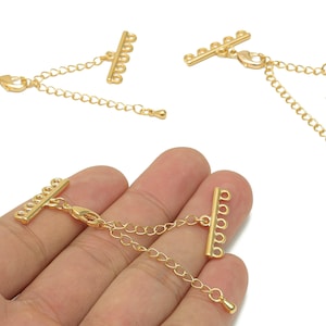 Puede incluir: Cierres de collar dorados con un diseño de barra y cadena. Los cierres tienen múltiples bucles para sujetar componentes de joyería. La imagen muestra los cierres en diferentes orientaciones, uno de ellos sostenido en una mano.