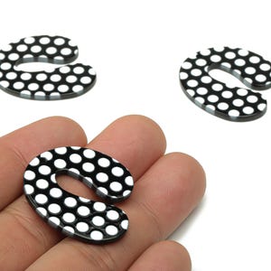 May include: Three black and white polka dot shaped charms. One charm is held in a hand, with two others scattered in the background. The charms are a glossy black with white polka dots. These charms are ideal for jewelry making.