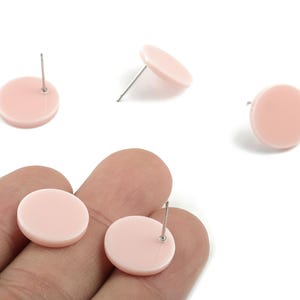 Puede incluir: Pendientes de botón redondos de color rosa pálido con postes plateados. Los pendientes se muestran sobre un fondo blanco, con algunos pendientes dispersos y otros sostenidos en una mano. Diseño simple y minimalista.