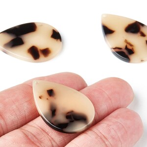 May include: Three teardrop-shaped beads with a tortoiseshell pattern. The beads have a cream-colored base with irregular dark brown spots. One bead is held in a hand, while the other two are on a white background.
