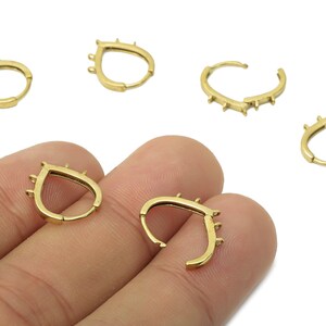 May include: Gold-colored hoop earrings with a teardrop design. Each earring features small, decorative spikes along the top edge. Several earrings are displayed, some open and some closed, against a white background.