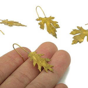 Puede incluir: Pendientes dorados con forma de hoja y cierre de gancho. Los pendientes tienen forma de hojas de arce y se muestran sobre un fondo blanco. Están hechos de un material metálico brillante.