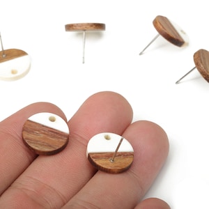 Pode incluir: Brincos redondos com design bicolor, combinando madeira e resina branca. Vários brincos são mostrados em fundo branco, alguns com os pinos visíveis.