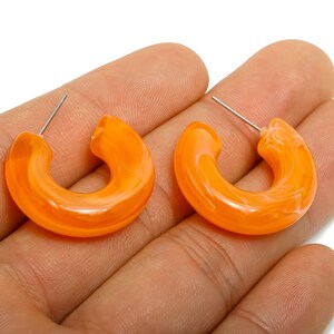 Puede incluir: Un par de pendientes de aro naranja con efecto veteado. Los pendientes son de color naranja brillante y translúcido y tienen un poste para orejas perforadas. Los pendientes se sostienen en la mano de una persona.
