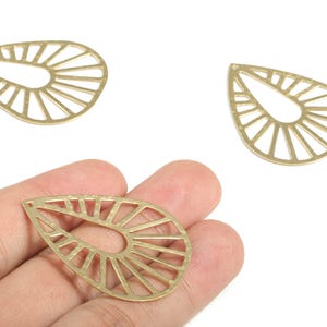 Puede incluir: Tres colgantes metálicos dorados en forma de lágrima con diseño de rayos de sol. Un colgante se sostiene en una mano. Los colgantes son probablemente para la fabricación de joyas u otros proyectos de artesanía.