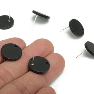 May include: Black, round stud earrings with silver posts. The earrings are made of a smooth, solid material and have a small hole near the top for the post. Several earrings are scattered on a white surface.
