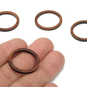 Puede incluir: Cuatro cuentas de madera en forma de anillo. Las cuentas son de color marrón cálido con un interior marrón oscuro. Una cuenta se sostiene en una mano. Las cuentas son probablemente para la fabricación de joyas o manualidades.