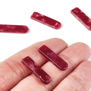 Puede incluir: Se muestran varias cuentas rectangulares de color granate con extremos redondeados. Las cuentas tienen un aspecto veteado con remolinos rojos y blancos más claros. Dos cuentas se sostienen en una mano, mientras que otras están sobre una superficie blanca.