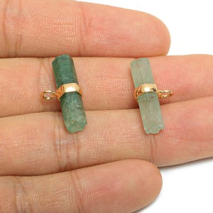 Puede incluir: Dos colgantes de cristal en bruto, verde y verde pálido, con detalles metálicos dorados. Los cristales son cilíndricos y tienen un lazo para colgar. Los colgantes se sostienen en una mano.