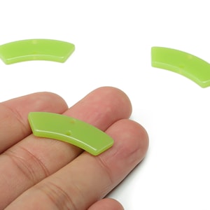 May include: Three lime green, curved, semi-circular shaped beads. One bead is held in a hand, while two others are positioned above. Each bead has a small hole for stringing. The beads are made of a glossy material.
