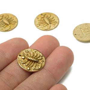 May include: Gold-coloured metal charms featuring a scorpion design. The round charms have a detailed embossed scorpion and star pattern. One charm is held in a hand, with three others scattered nearby.