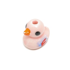 May include: A pink ceramic duck bead with black eyes, an orange beak, and blue and red details.
