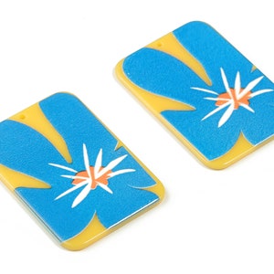 May include: Two yellow and blue rectangular earrings with a flower design. The flower is blue with a white center and orange accents.