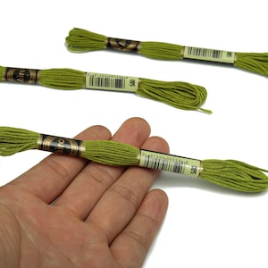 Puede incluir: Tres madejas de hilo de bordar verde. Una se sostiene en una mano. El hilo está enrollado alrededor de un tubo de cartón con una etiqueta que incluye el número 580. El hilo es de un color verde brillante y primaveral.