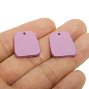 May include: Two lavender-colored, rectangular pendants with rounded corners and a small hole at the top. The pendants are smooth and matte, and are held in a person's hand against a white background. These pendants are suitable for jewelry making.