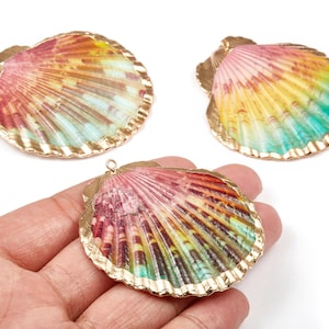 Puede incluir: Tres colgantes de concha coloridos con bordes dorados. Las conchas tienen un degradado de colores, incluyendo rosa, amarillo y azul. Una concha se sostiene en una mano. Estos colgantes son ideales para la fabricación de joyas.