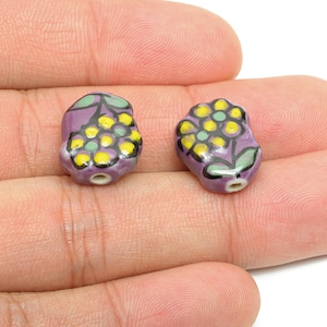 May include: Two ceramic beads with a floral design. The beads are purple with yellow and green flower details and black outlines. Each bead has a small hole for stringing.