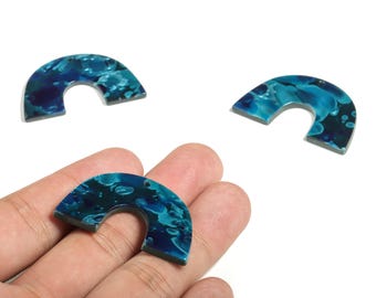 May include: Three arch-shaped pendants with a marbled blue and teal design. One pendant is held in a hand. The pendants have a glossy finish and are likely made of resin or acrylic. These are suitable for jewelry making.