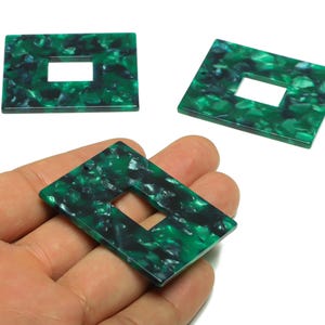 May include: Three green and black rectangular acrylic pendants with a central rectangular cutout. One pendant is held in a hand. The pendants have a marbled pattern, suitable for jewellery making or crafting.