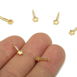 May include: Gold-colored star charms with a dangling design. Each charm features a five-pointed star and a linear section adorned with small, clear crystals. The charms are small, with a loop for attachment, and are displayed against a white background.