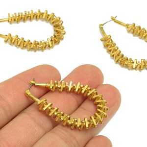May include: Gold-colored hoop earrings with a unique design. The earrings have a series of small, rectangular bars arranged around the hoop. One earring is held in a hand, and two others are displayed on a white background.