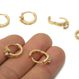 May include: Gold-colored hoop earrings with a small, clear gemstone and additional small clear stones. The earrings are shown in multiple views, with some held in a hand.