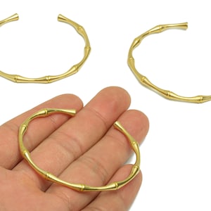 May include: Gold-coloured bamboo-style bracelets. The open cuff design features a series of connected segments, mimicking the look of bamboo stalks. One bracelet is held in a hand, while two others are displayed separately.