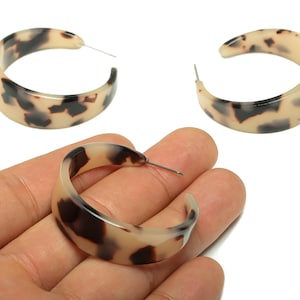 May include: Three tortoise shell hoop earrings. The earrings are a light beige color with dark brown spots. One earring is held in a hand. The earrings have a post for pierced ears.