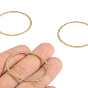May include: Three gold-colored, open circle jewelry findings. One is held in a hand, while two others are on a white surface. These circular components are used in jewelry making for earrings, pendants, or other designs.