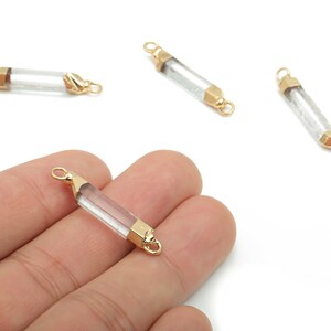 May include: Clear, cylindrical crystal beads with gold-colored metal caps and loops. The beads are displayed on a white background, with one held in a hand. These beads are suitable for jewelry making.