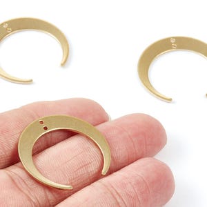 May include: Three gold-coloured crescent moon charms with two small holes at the top for stringing. One charm is held in a hand. The charms are made of metal and are on a white background.