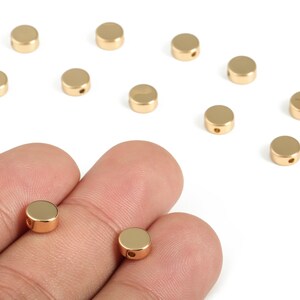 May include: Gold-coloured, flat, round beads with a hole through the centre. Several beads are scattered on a white surface, with two held in a hand. These beads are likely used for jewellery making or crafting.
