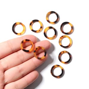 May include: A collection of circular beads with a tortoiseshell pattern. The beads are a mix of amber and dark brown, with a glossy finish. Some beads are held in a hand, while others are scattered on a white surface. These beads are suitable for jewelry making.