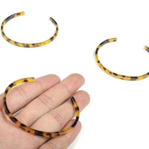 Puede incluir: Tres brazaletes abiertos con un patrón de carey. Los brazaletes son amarillos, marrones y negros. Un brazalete se sostiene en una mano, mientras que los otros dos están sobre un fondo blanco.