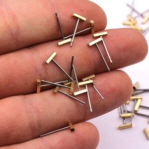 May include: A collection of small, minimalist stud earrings. Each earring features a gold-colored rectangular bar attached to a silver-colored post. The earrings are scattered across a hand, showcasing their simple design.