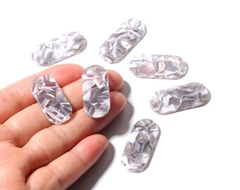 May include: A collection of seven oval-shaped beads with a marbled, pearlescent design in shades of gray and white. The beads are shown against a white background, with one held in a hand.