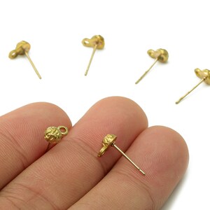 May include: Gold-colored stud earrings with a textured, heart-shaped design. The earrings have a post for pierced ears. Several earrings are scattered on a white background.