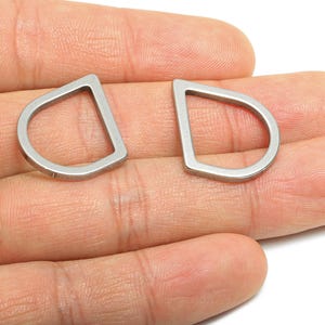 May include: Two silver-colored, D-shaped metal findings. Each finding has a flat top and a rounded bottom, with a consistent width throughout. The items are held in a hand, showcasing their size and design. These findings are likely used in jewelry or craft projects.