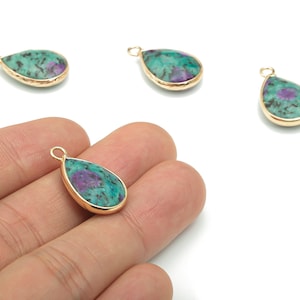 Peut inclure: Quatre pendentifs en forme de goutte d'eau avec une lunette dorée. Les pendentifs présentent une pierre turquoise et violette. Un pendentif est tenu dans une main. Le fond est blanc.