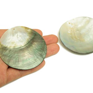 May include: Two iridescent, round, mother-of-pearl shell pendants. One is held in a hand, displaying a mix of green, white, and gold hues. The other shell pendant is visible to the right, showcasing similar colours and a natural, organic texture.