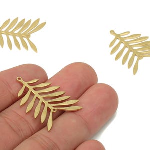 May include: Gold-colored leaf charms with a detailed fern design. The charms are shown in a hand, with two additional charms in the background. These charms are ideal for jewelry making and crafting projects.