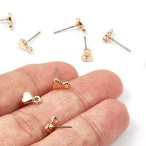 May include: Gold-coloured heart-shaped stud earrings with a post. Several earrings are scattered on a white surface. The earrings are small and delicate, with a smooth, polished finish. The earrings are shown in a person's hand.