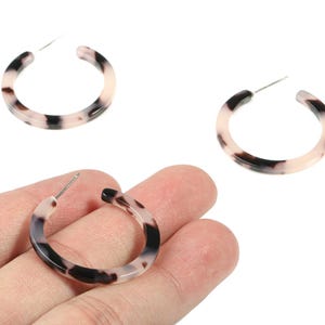 May include: Three hoop earrings with a tortoiseshell pattern. The earrings are a mix of black, white, and tan colors. The earrings are held in a hand against a white background. The earrings are a jewelry item.