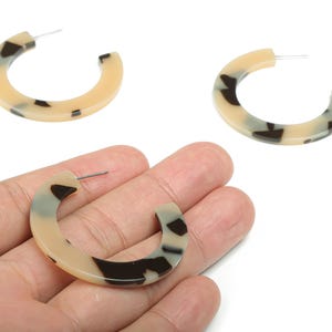 May include: Tortoiseshell-patterned hoop earrings. The earrings have a C-shape with a post for pierced ears. The color pattern includes tan, black, and gray spots. The earrings are shown against a white background.
