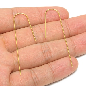 May include: A pair of gold-coloured, minimalist earrings. Each earring features a long, straight post that curves into a U-shape at the top. The earrings are displayed on a hand, highlighting their delicate design. These earrings are a simple yet elegant accessory.