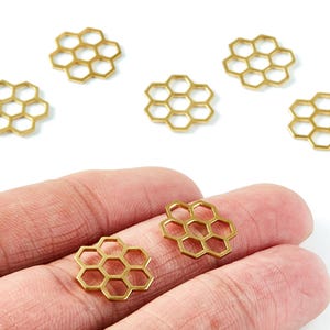 Puede incluir: Encantos dorados en forma de panal. Los encantos están dispuestos sobre un fondo blanco, con dos sostenidos en una mano. Estos encantos probablemente se utilizan para la fabricación de joyas u otros proyectos de artesanía.