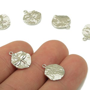 Puede incluir: Encantos de metal plateado con una superficie texturizada y un diseño de libélula. Los encantos tienen un pequeño lazo para sujetarlos a joyas u otras manualidades. Varios encantos son visibles, algunos se sostienen en una mano.