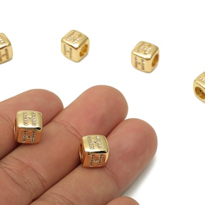 Peut inclure: Perles dorées en forme de cube avec la lettre "H" sur chaque face, ornées de petits cristaux transparents. Les perles sont présentées sur un fond blanc.