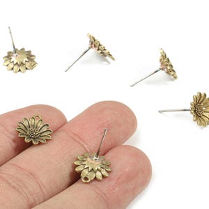 May include: Antique gold-colored daisy stud earrings. The earrings feature a detailed floral design with a post for pierced ears. The image shows several earrings scattered on a white surface.